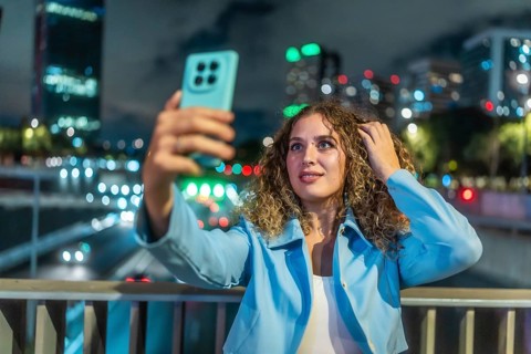 Young woman with curly hair taking a selfie with her mobile phone on a city bridge at night,