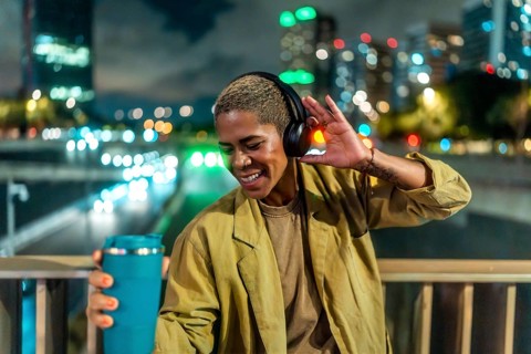Woman listening to music with headphones and holding a travel mug, smiling brightly while standing