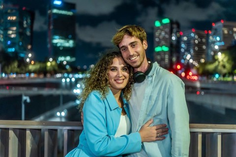 Young couple embracing and smiling at the camera outdoors on a bridge, with illuminated city