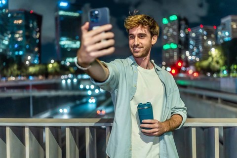 Young man standing on a bridge, holding a reusable cup and smiling while taking a selfie with a