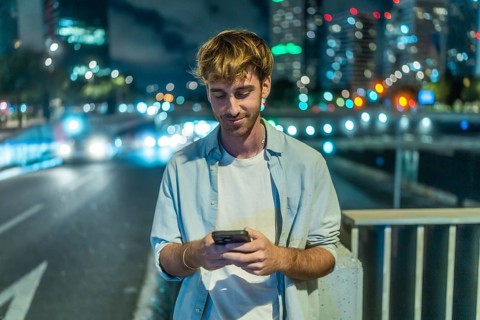 Young man standing on a city bridge at night. Engaging with a smartphone. With blurred city lights