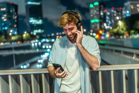 Young man smiling and listening to music through wireless headphones, holding a smartphone while