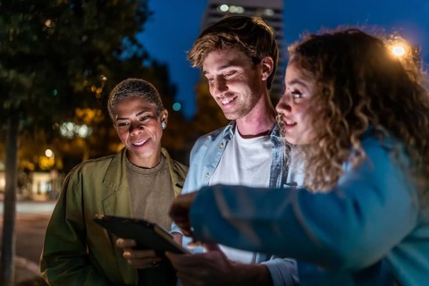 Three diverse friends smiling and interacting outdoors at night, using a digital tablet to share