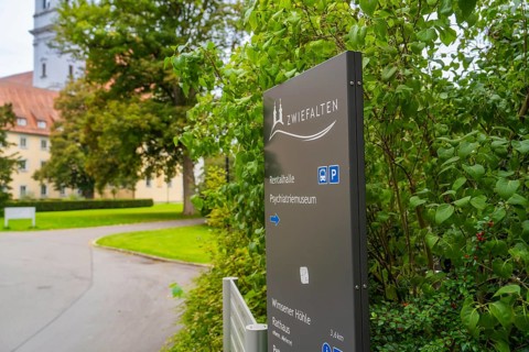 A sign with the reference to Zwiefalten stands against a background of green plants and a building,