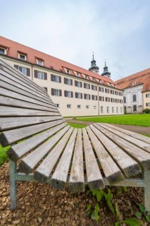 A wooden park bench in the foreground with a monastery building in the background under a cloudy