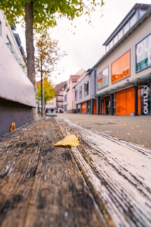 A quiet shopping street with modern buildings, autumn leaves and a wooden bench in the foreground,