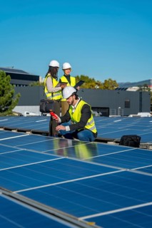 Engineers in hard hats and safety vests are examining solar panels on a rooftop, checking