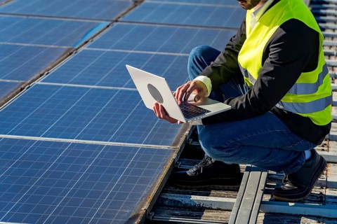 Engineer wearing a high visibility vest and crouching on a rooftop, using a laptop to monitor and