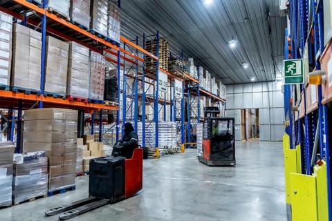 Workers are managing inventory with forklifts, navigating between tall shelving units stacked with