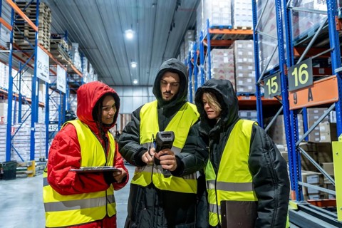 Three warehouse workers in protective clothing and high visibility vests are collaborating while