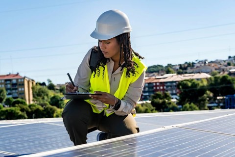Black woman engineer performing an inspection of photovoltaic solar panels on a rooftop, writing