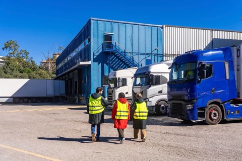 Three workers wearing safety vests walking towards a blue industrial building with multiple semi