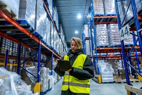 Woman worker in protective clothing and high visibility vest checking goods on shelves with a