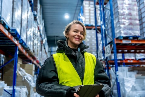 Woman worker smiling in safety vest, holding a tablet, standing in a large cold storage warehouse