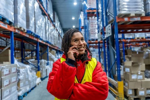 Woman warehouse worker wearing warm clothing and a safety vest, smiling while having a phone