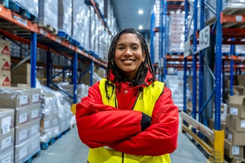 Smiling young woman standing with arms crossed in a brightly lit cold storage warehouse. Wearing
