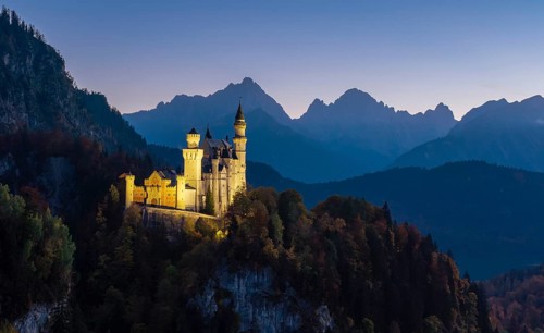 Illuminated Neuschwanstein Castle in front of a rugged mountain panorama, nestled in a dense forest
