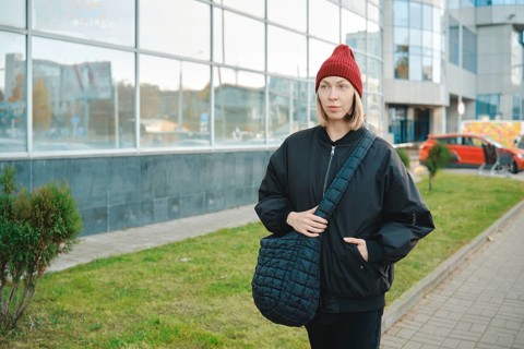 A woman wearing a black jacket and red beanie walks along a modern glass building. She holds a
