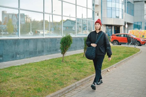 A woman strolls along a wide walkway surrounded by a mix of urban buildings and greenery. She wears