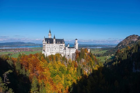 The magnificent Neuschwanstein Castle is enthroned over colorful forests with a bright blue sky,