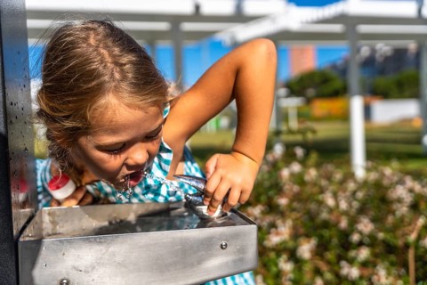 Little girl bending to drink cool water from a stainless steel park fountain on a sunny summer day,