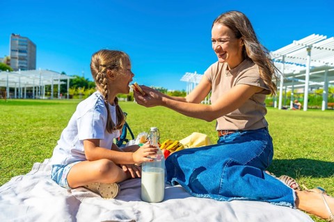 Young mother feeding her little daughter a sandwich during a sunny summer picnic on a blanket in