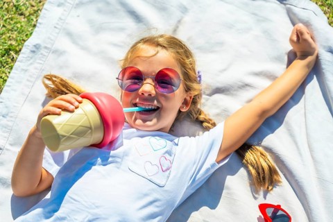 Young girl lying on a blanket in the grass on a sunny day, laughing while drinking from an ice