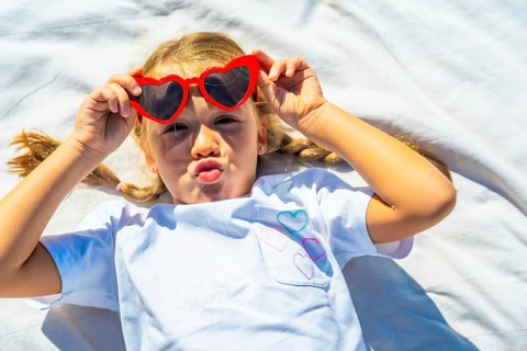 Young blonde girl lying on a white blanket making a fun kiss face while wearing red heart shaped