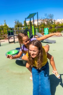 Mother gives daughter a piggyback at a sunny playground, both laughing and smiling as they enjoy