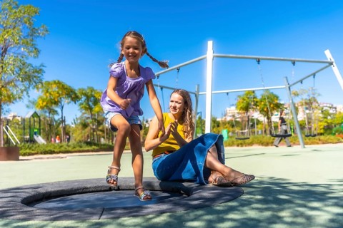 Daughter jumps on a round trampoline at a sunny park playground while mother claps nearby, sharing