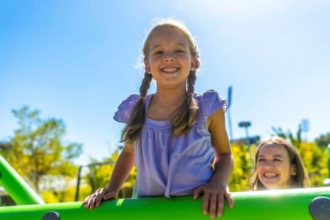 Young girl with pigtails smiling at the camera while her mother watches from playground equipment