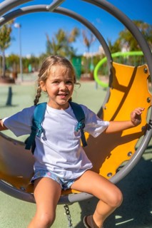 Happy little girl with a backpack smiles broadly while sitting on a swing in a vibrant outdoor
