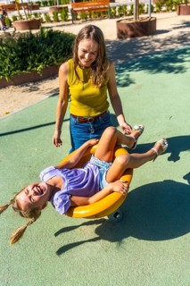 Mother standing, smiling, and gently pushing her happy daughter on a yellow spinning toy, both