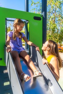 Mother encouraging her smiling daughter on a sunny park slide, sharing a playful bonding moment of
