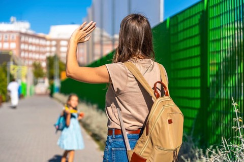 Mother waving goodbye, watching her young daughter with a backpack walk independently towards