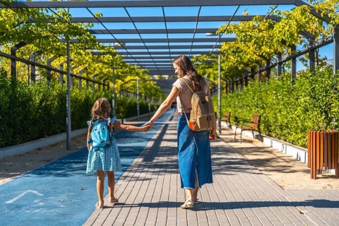Mother and daughter walking along an urban path, holding hands and carrying backpacks on a sunny