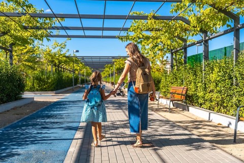 Mother and daughter, wearing backpacks, walking along a sunny pathway lined with green trees and