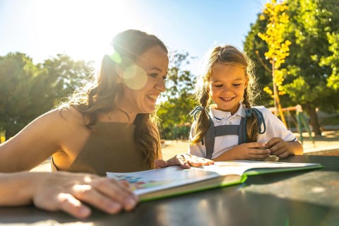 Mother and daughter smiling together at an outdoor schoolyard table, reading a book and bonding