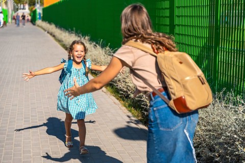 Child daughter with backpack runs across schoolyard to joyfully hug her mother at pickup,