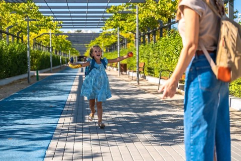 Excited schoolgirl with backpack runs toward her mother in a sunny park, holding a banana after