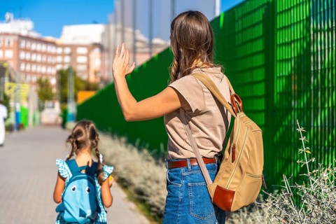 Mother waving with her hand to a small daughter carrying a backpack and walking away towards