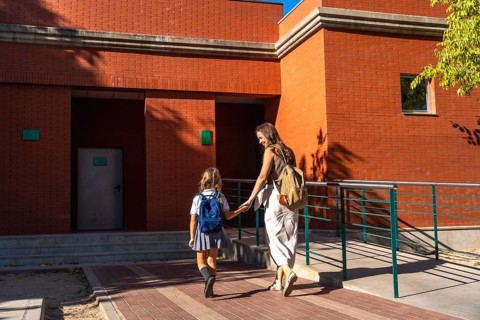 Mother holding hands with her daughter, both walking together towards a brick school building on a