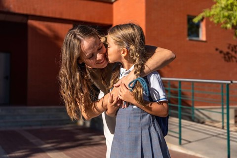Mother embraces and kisses her young daughter in school uniform at the school entrance on the first
