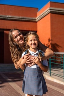 Mother and daughter smiling while standing together on a sunny day at school with the elementary