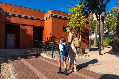 Mother and daughter smiling as they walk toward a red brick school building with backpacks,