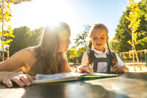 Mother and daughter bonding over reading a book together, sitting at a table outdoors in a bright