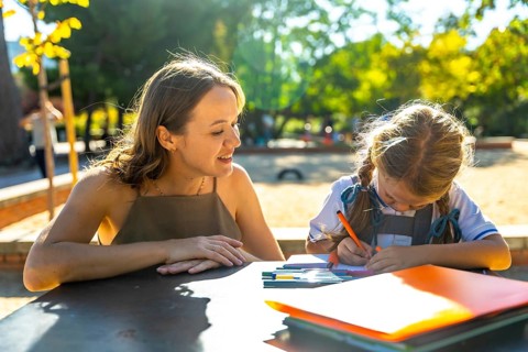 Mother watching her daughter drawing and doing homework at an outdoor table at school, enjoying a