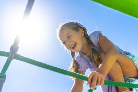 Happy young girl laughing with joy while actively climbing a green rope net structure outside on a