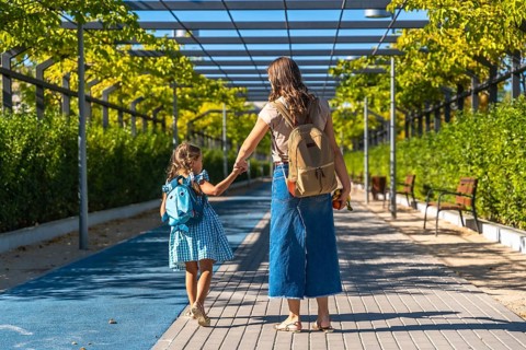 Mother and daughter holding hands and walking together along a paved path, carrying backpacks and