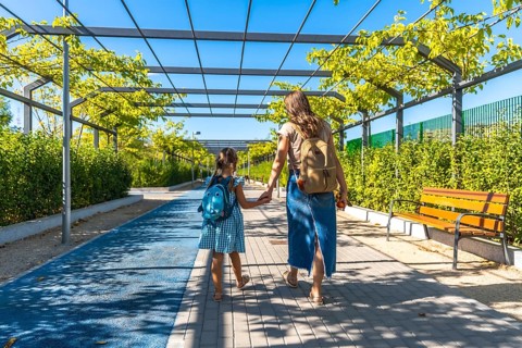 Mother and daughter walk hand in hand along an urban park path with backpacks, heading to school on